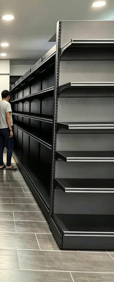 A newly installed run of black gondola shelving in a mini market, with a shopper at the far end of the aisle.